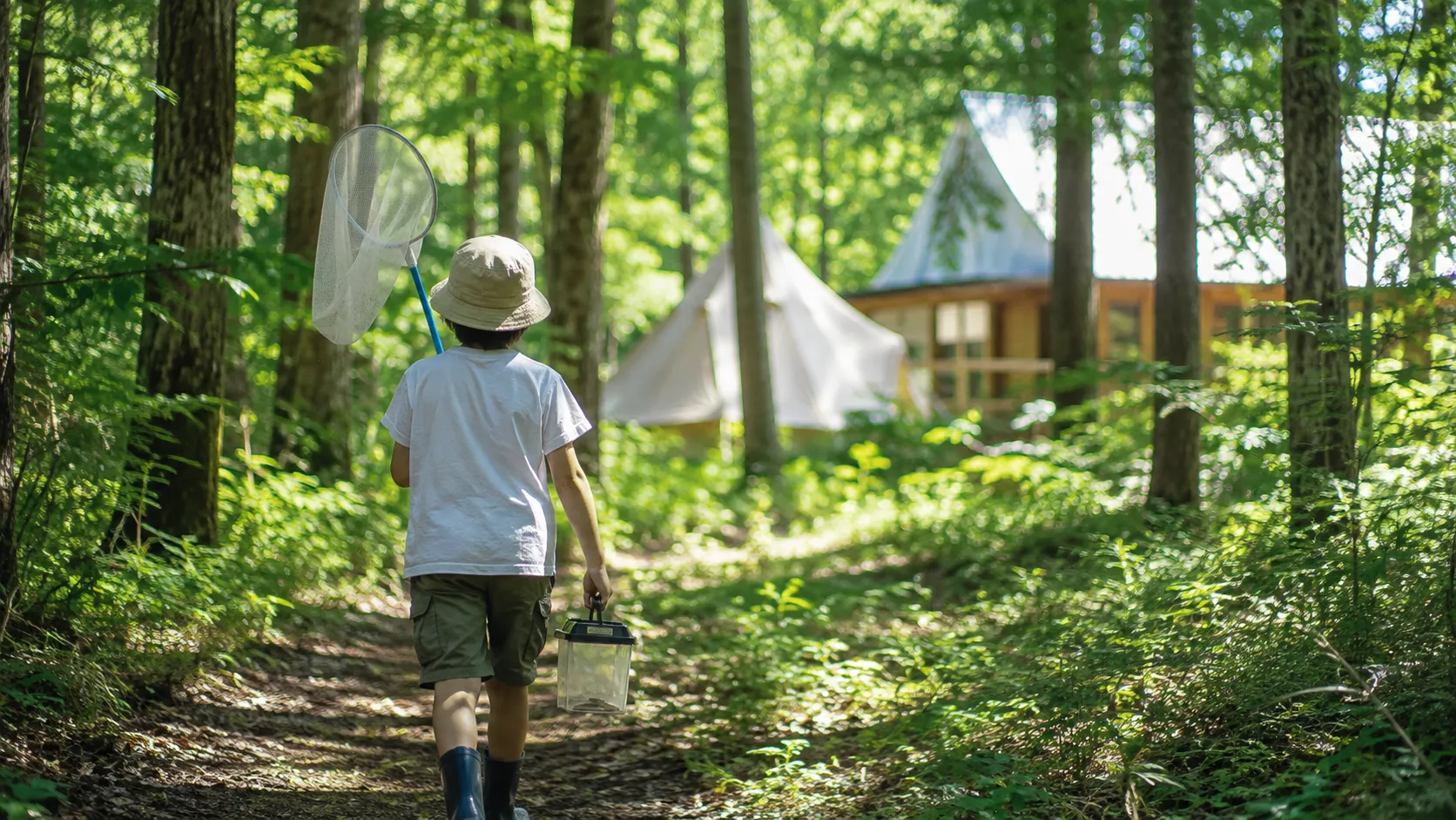 A child walking a sunlit forest path near glamping tents, carrying a Japanese insect net and bug cage